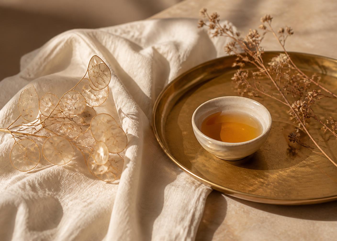 Brass tray with a ceramic bowl of golden oil and dried botanicals on ivory linen